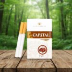 บุหรี่ Capital Coffee ซองสีขาวแถบน้ำตาลลายเมล็ดกาแฟ วางบนโต๊ะไม้ฉากหลังป่าสีเขียว